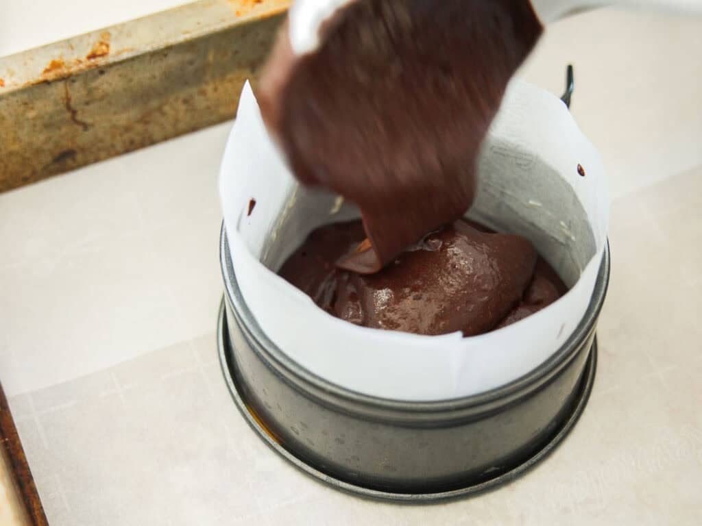 Chocolate cake batter is being poured into a round springform pan lined with parchment paper, placed on a baking tray.