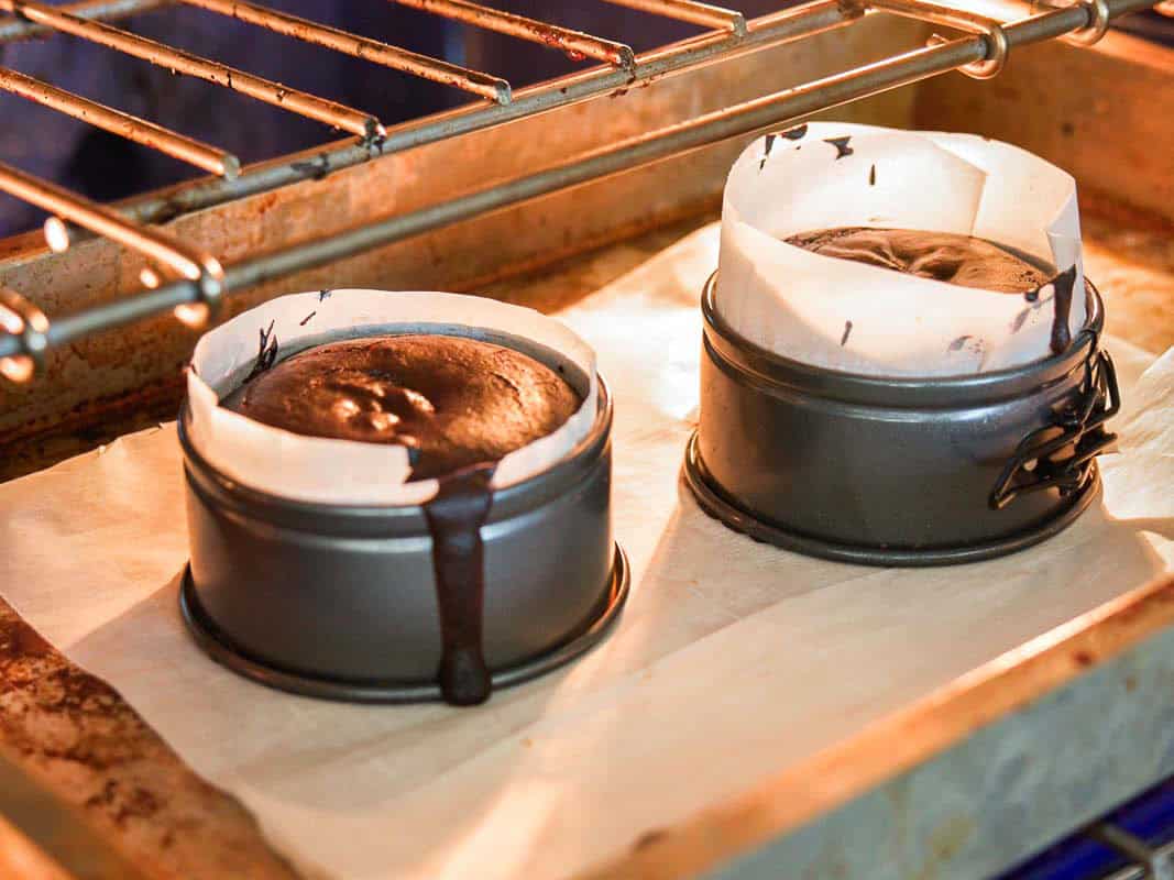 Two small round cake pans lined with parchment paper, filled with chocolate cake batter, are baking in an oven. One pan has batter overflowing down the side onto the baking sheet below.