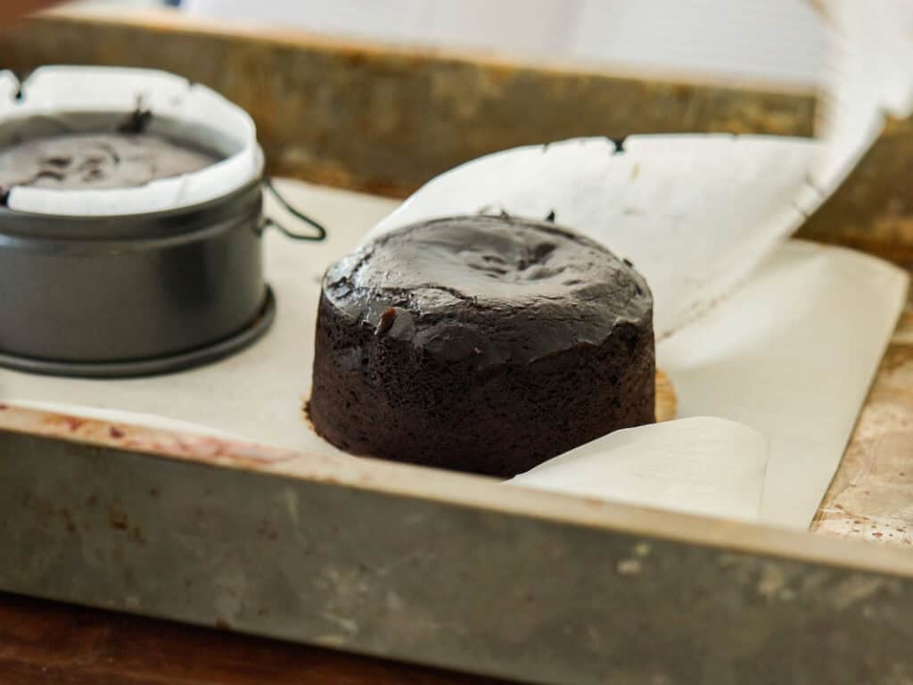 A freshly baked round chocolate cake sits on parchment paper in a metal baking tray, with another cake still in a round springform pan nearby.