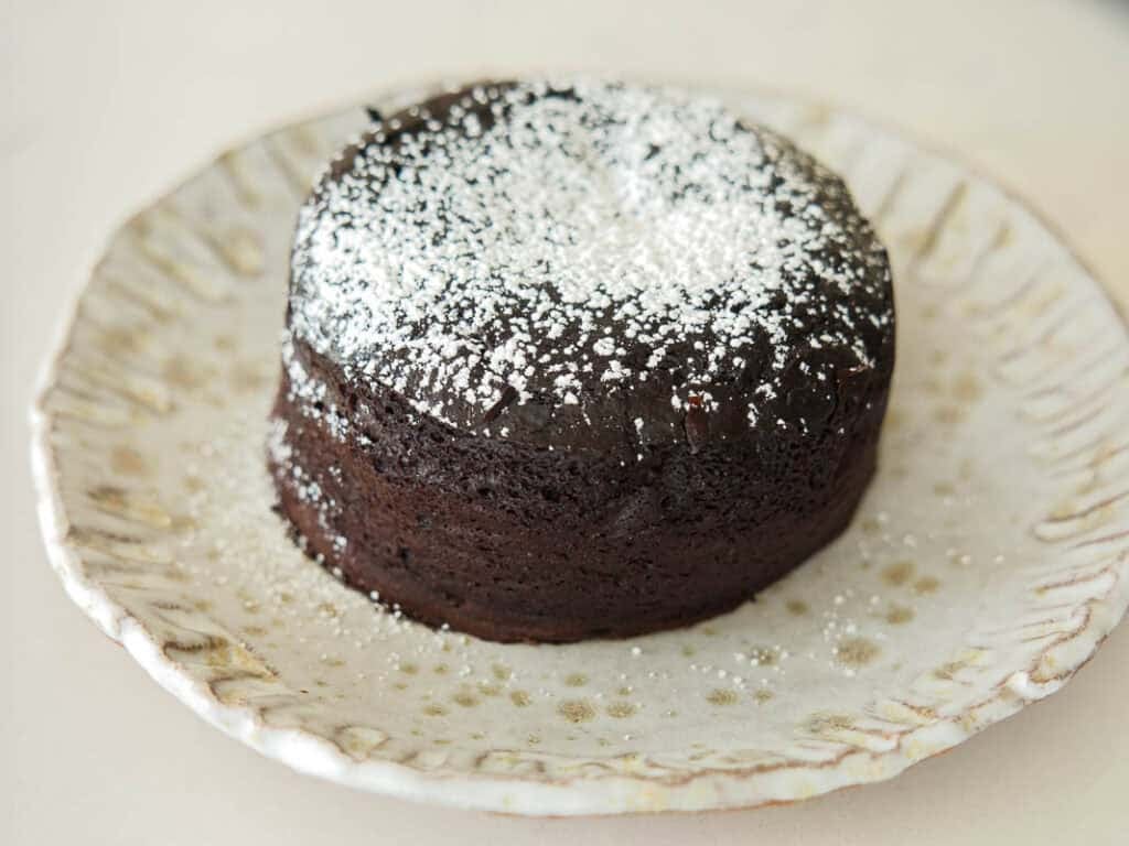 A round vegan chocolate lava cake topped with powdered sugar sits on a decorative ceramic plate with a scalloped edge.