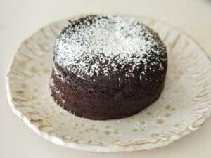 A round vegan chocolate lava cake topped with powdered sugar sits on a decorative ceramic plate with a scalloped edge.