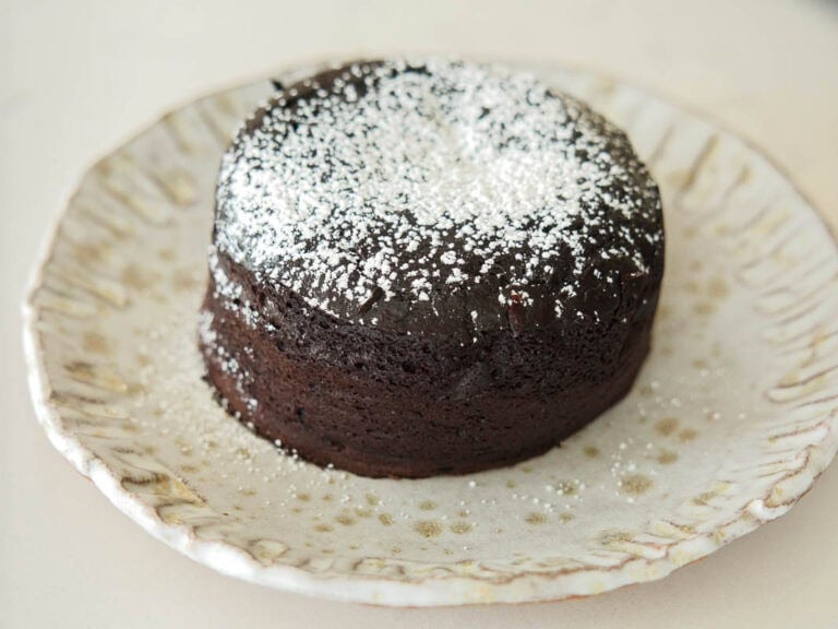Recipes 7 A round vegan chocolate lava cake topped with powdered sugar sits on a decorative ceramic plate with a scalloped edge.