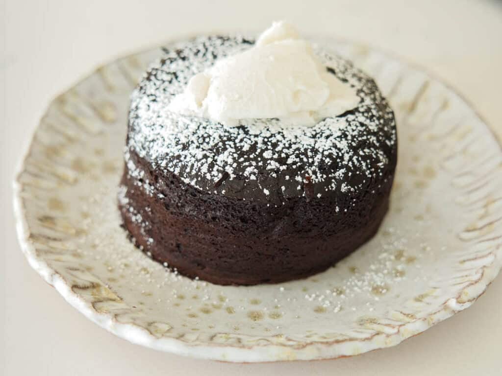 A round vegan chocolate lava cake dusted with powdered sugar, topped with a dollop of whipped cream, served on a rustic, white ceramic plate.