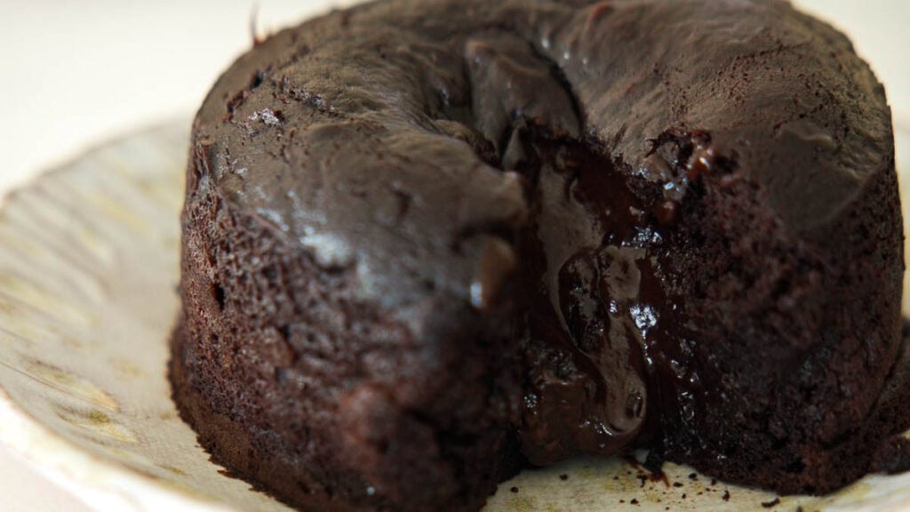 A close-up of a vegan chocolate lava cake on a plate, with rich, melted chocolate oozing from the center of the moist, dark cake.