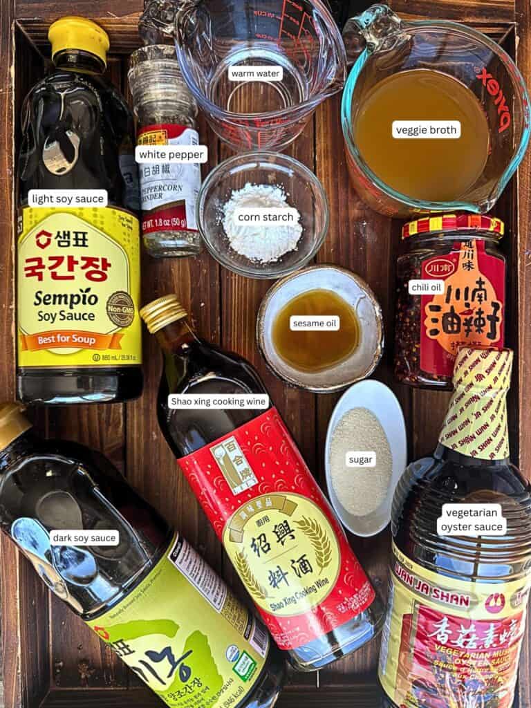 Easy & Healthy Lo Mein Vegan Recipe: Done in Minutes. 21 A wooden tray displays ingredients for Asian cooking, including light soy sauce, Shao Xing cooking wine, dark soy sauce, white pepper, cornstarch, sesame oil, sugar, warm water, veggie broth, vegetarian oyster sauce, and chili oil.