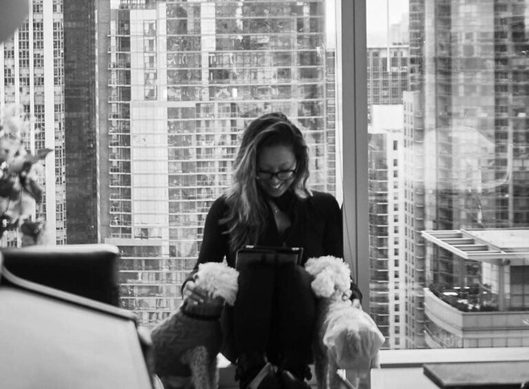 Joanne sits by a large window in a city high-rise, smiling at a tablet on her lap, while two small dogs rest their heads on her knees. Tall buildings are visible in the background. Black and white photo.