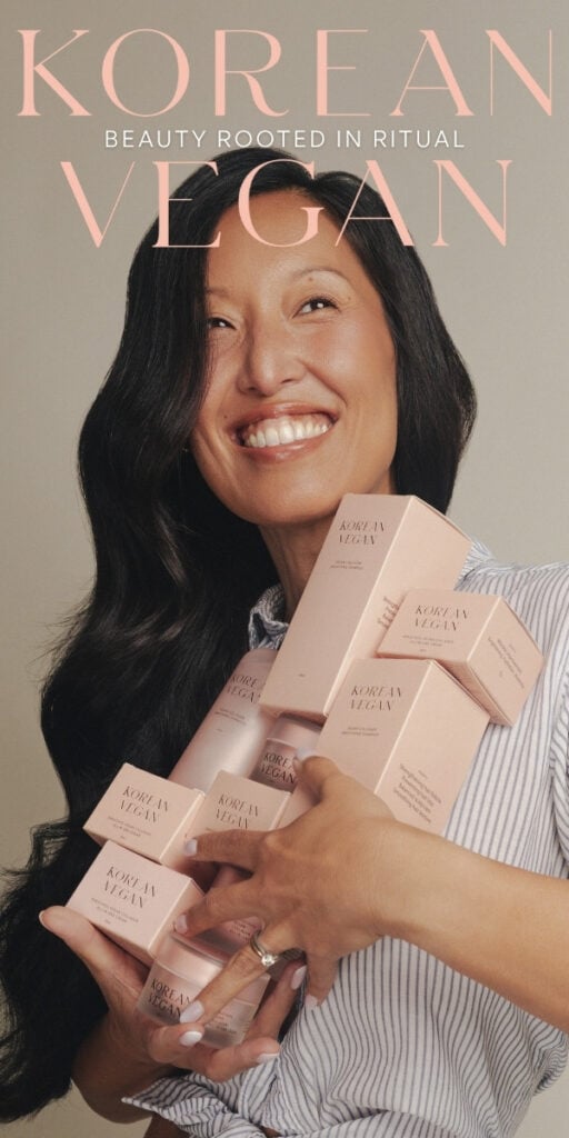 Smiling woman with long dark hair holds several pink boxes labeled Korean Vegan against a beige background. Text above reads “Korean Vegan: Beauty Rooted in Ritual.”.