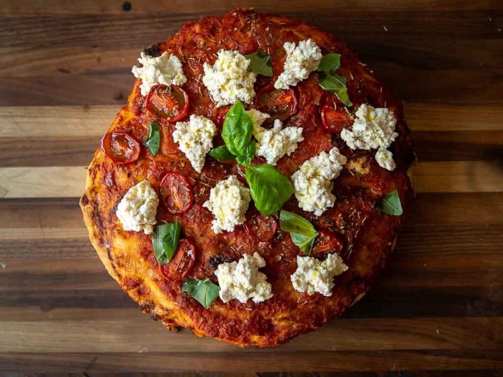 A round focaccia rossa topped with tomato sauce, roasted cherry tomatoes, dollops of ricotta cheese, and fresh basil leaves, placed on a wooden surface.
