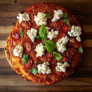 A homemade focaccia rossa topped with tomato sauce, roasted tomato slices, dollops of ricotta cheese, and fresh basil leaves, shown on a wooden surface.