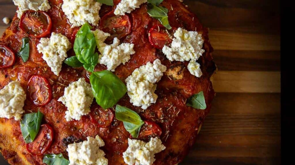 A close-up of a rustic focaccia rossa opped with tomato sauce, cherry tomato slices, dollops of ricotta cheese, fresh basil leaves, and herbs on a wooden surface.