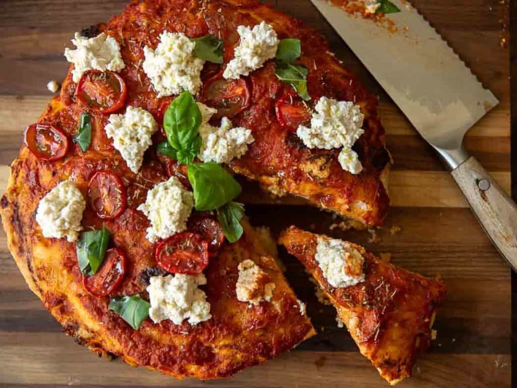 A focaccia rossa topped with tomato sauce, fresh basil leaves, sliced cherry tomatoes, and dollops of vegan ricotta cheese on a wooden board. A slice is being separated with a large knife.