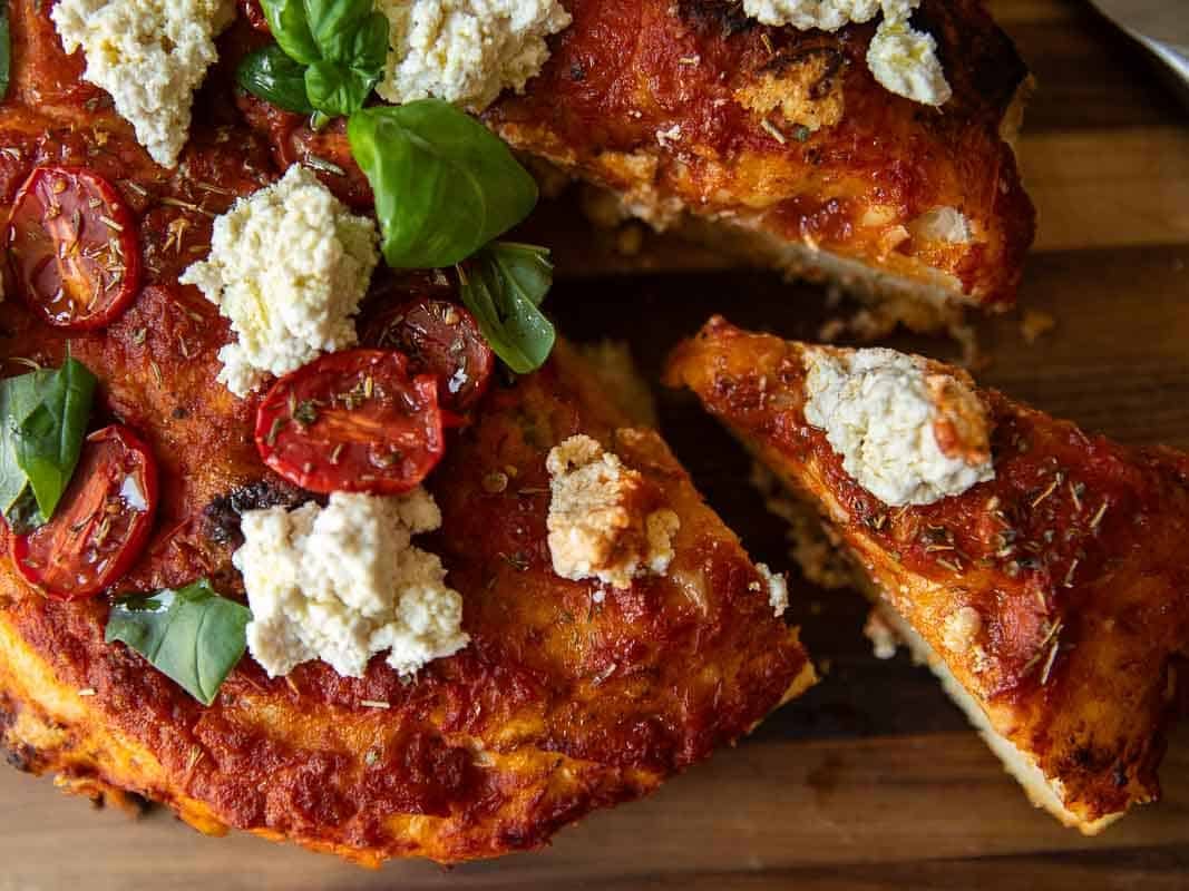 A close-up of a focaccia rossa topped with tomato sauce, roasted cherry tomatoes, dollops of vegan ricotta, and fresh basil leaves on a wooden surface. One slice is slightly pulled away from the pizza.