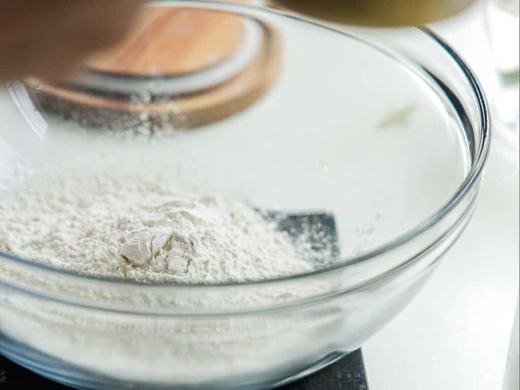 A glass mixing bowl containing a mound of white flour sits on a black surface, with a blurred wooden lid and hand visible in the background.
