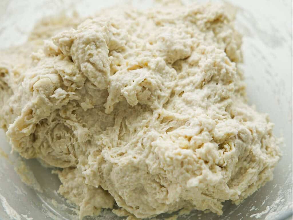 A close-up of raw bread dough, showing a rough, sticky texture with visible lumps and a pale off-white color. The dough appears to be freshly mixed and unbaked.