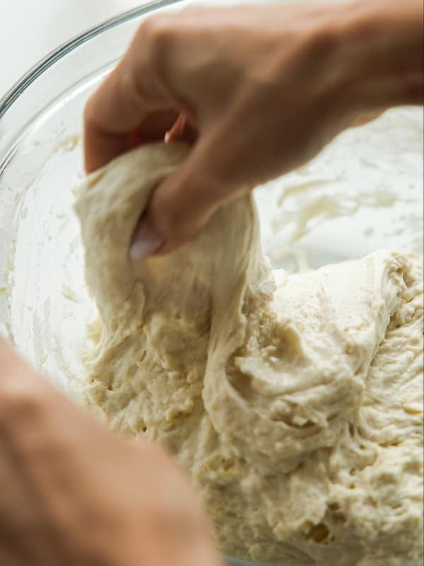 Close-up of hands stretching and folding soft bread dough in a glass bowl. The dough appears sticky and airy, indicating it is in the mixing or kneading stage of preparation.