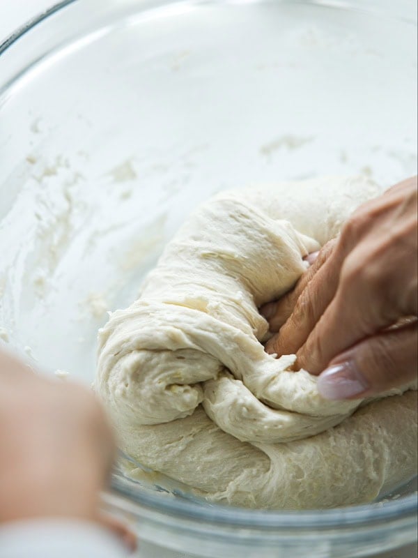 A close-up of hands kneading dough in a clear glass bowl, with soft, stretchy dough being pressed and folded.