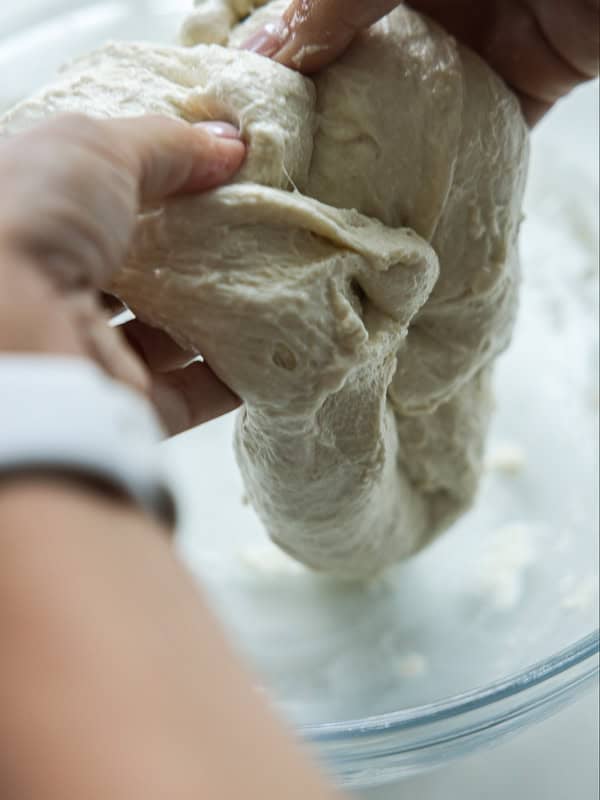 Close-up of hands stretching and folding sticky bread dough in a glass bowl, preparing it for baking.