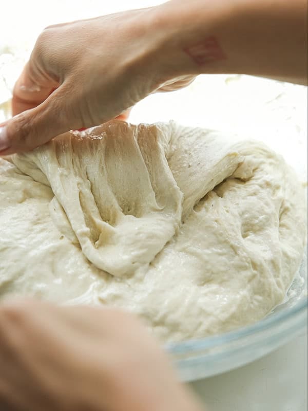 A close-up of hands stretching and folding a soft, sticky dough in a glass bowl, preparing it for baking.