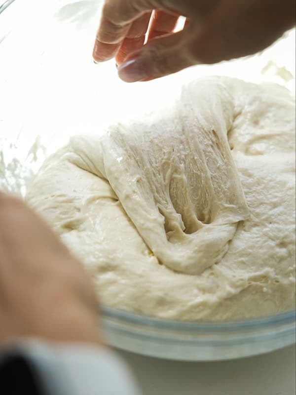 A close-up of hands stretching and folding soft, risen bread dough in a glass bowl.