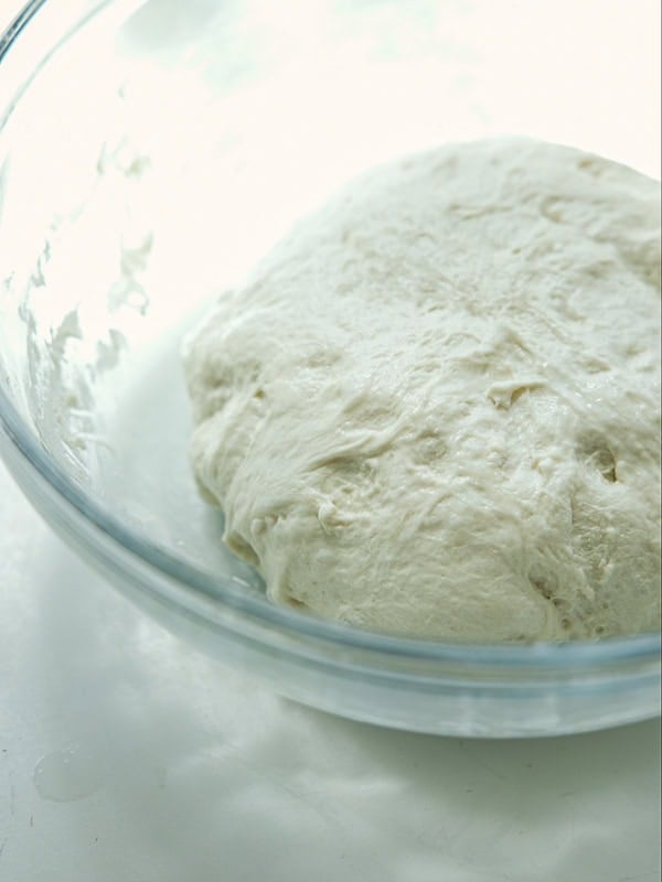 A close-up of a ball of risen dough in a clear glass bowl on a white surface, ready for baking.