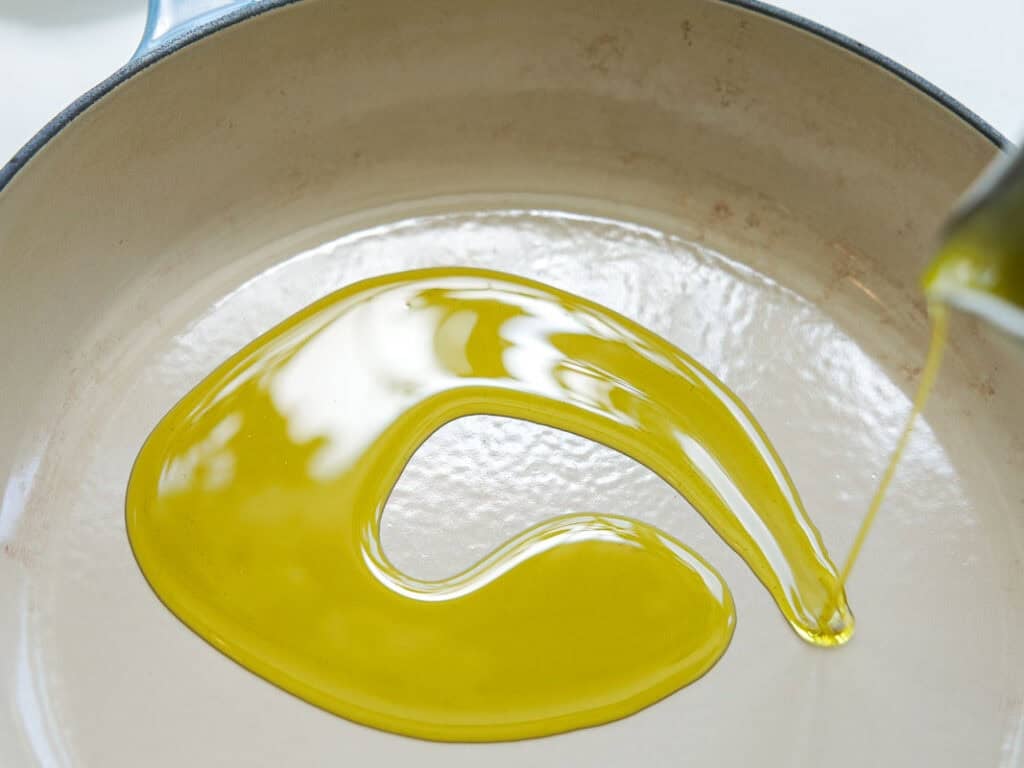 Olive oil being poured into a light-colored, empty skillet, forming a glossy swirl across the pan’s surface.