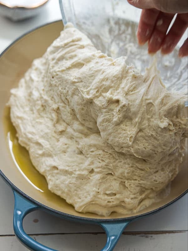 A hand transfers soft, airy bread dough from a glass bowl into a blue enameled baking dish with a layer of olive oil on the bottom.