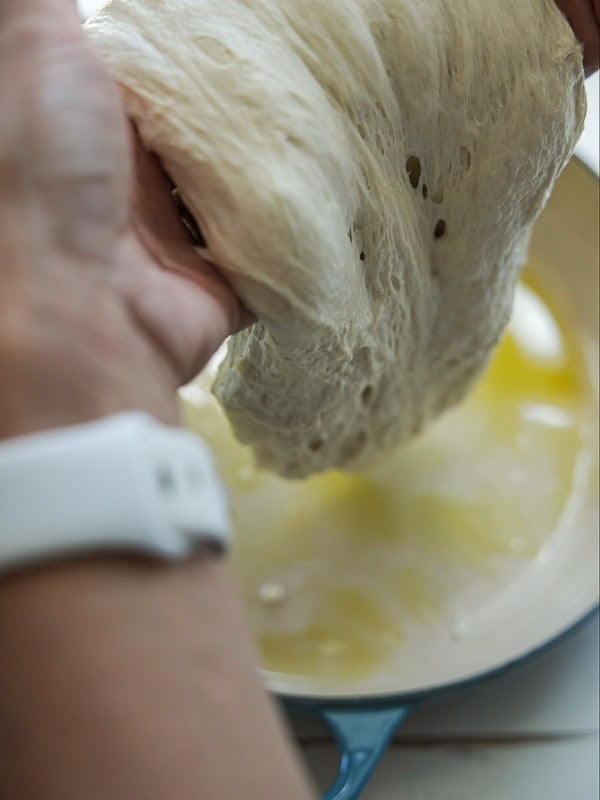 A person with a white watch stretches soft, airy bread dough over a blue dish filled with olive oil.