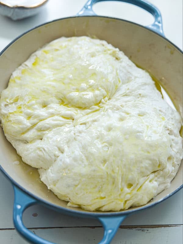 A round blue baking dish filled with risen, unbaked focaccia dough, glistening with olive oil on the surface.