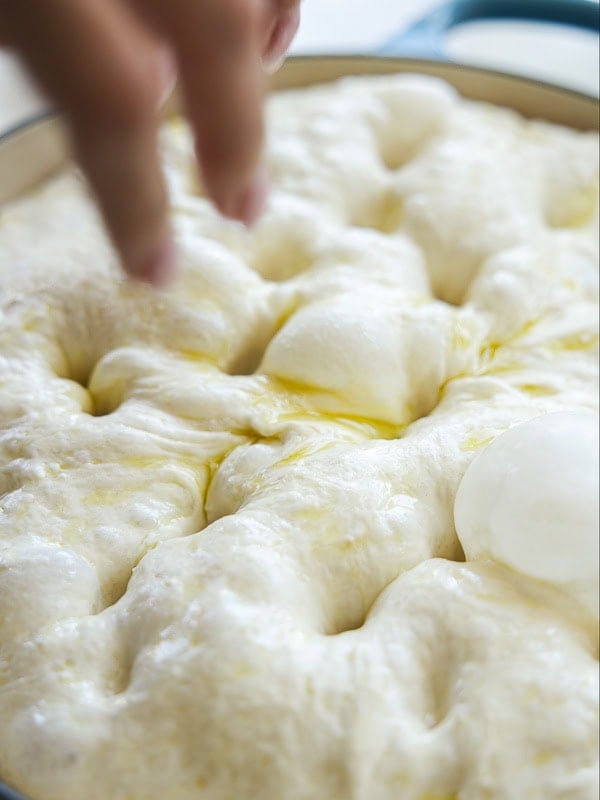 A close-up of a hand pressing fingers into soft, risen focaccia dough in a pan, with pools of olive oil visible on the surface.