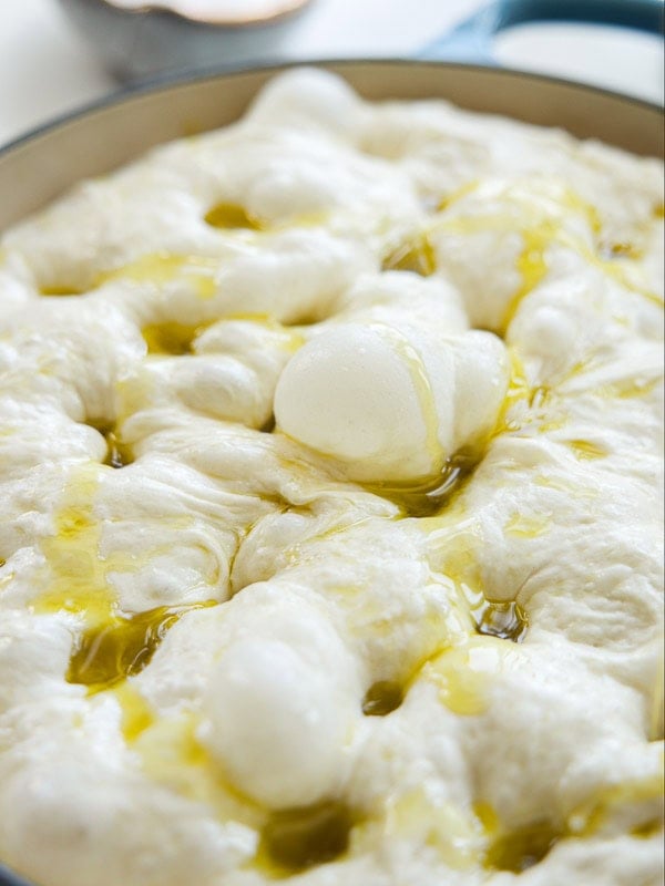 Close-up of soft, bubbly focaccia dough with dimples, drizzled with olive oil, in a round baking pan.