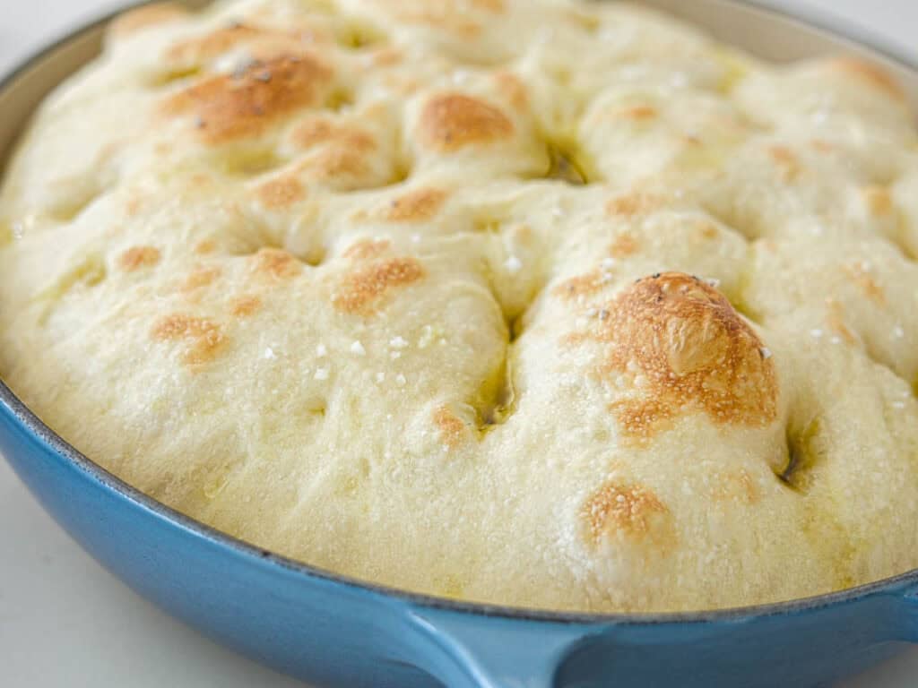 Golden, bubbly focaccia bread with a crisp top and dimpled surface, baked in a blue round pan and drizzled with olive oil.