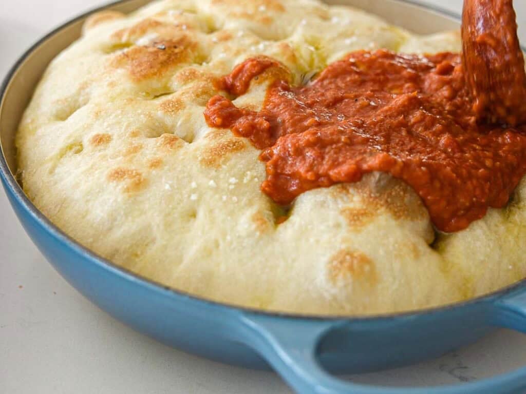 A close-up of tomato sauce being poured over freshly baked focaccia bread in a blue baking dish. The bread is golden and dimpled, and the sauce is thick and red.