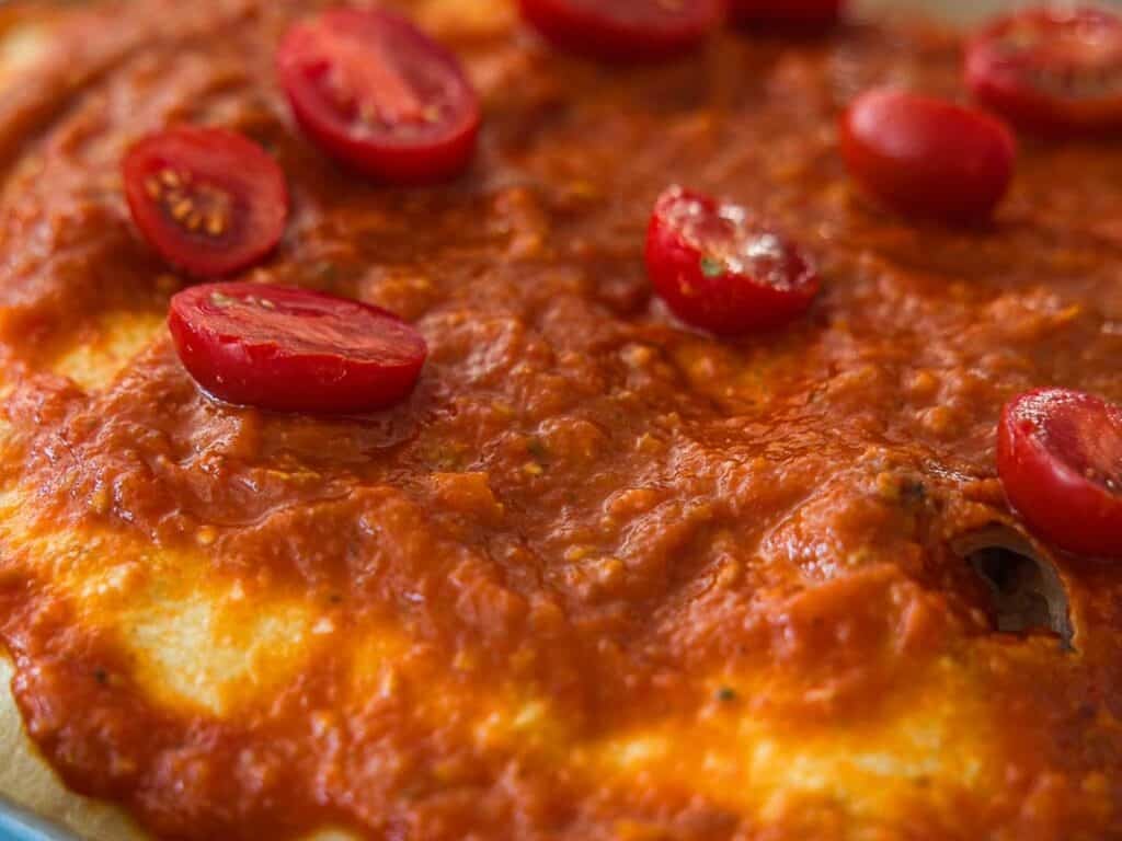 Close-up of a focaccia topped with tomato sauce and sliced cherry tomatoes, showing a golden-brown crust and a textured tomato surface.