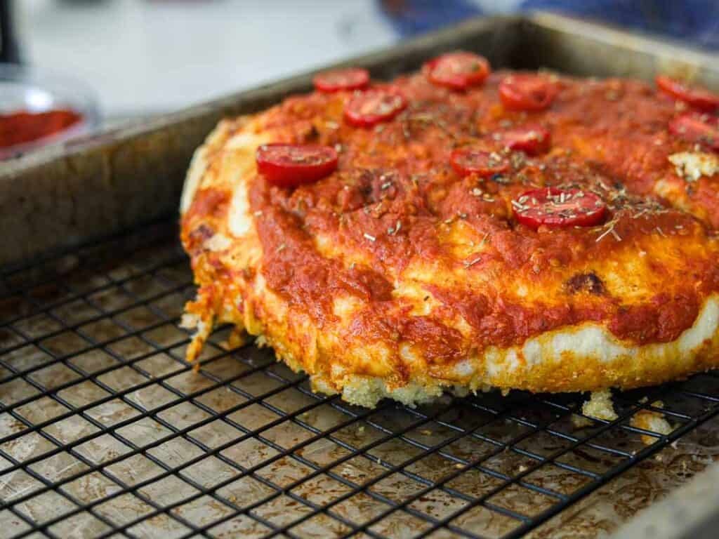 A thick, golden-brown focaccia rossa topped with tomato sauce, sliced cherry tomatoes, and dried herbs sits on a cooling rack over a baking sheet.