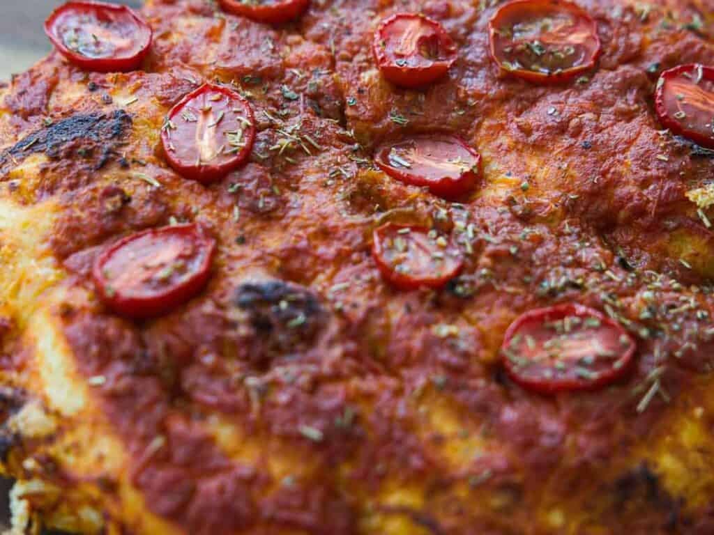 Close-up of a baked focaccia topped with tomato sauce, halved cherry tomatoes, and a sprinkle of dried herbs. The crust appears golden and slightly crispy.