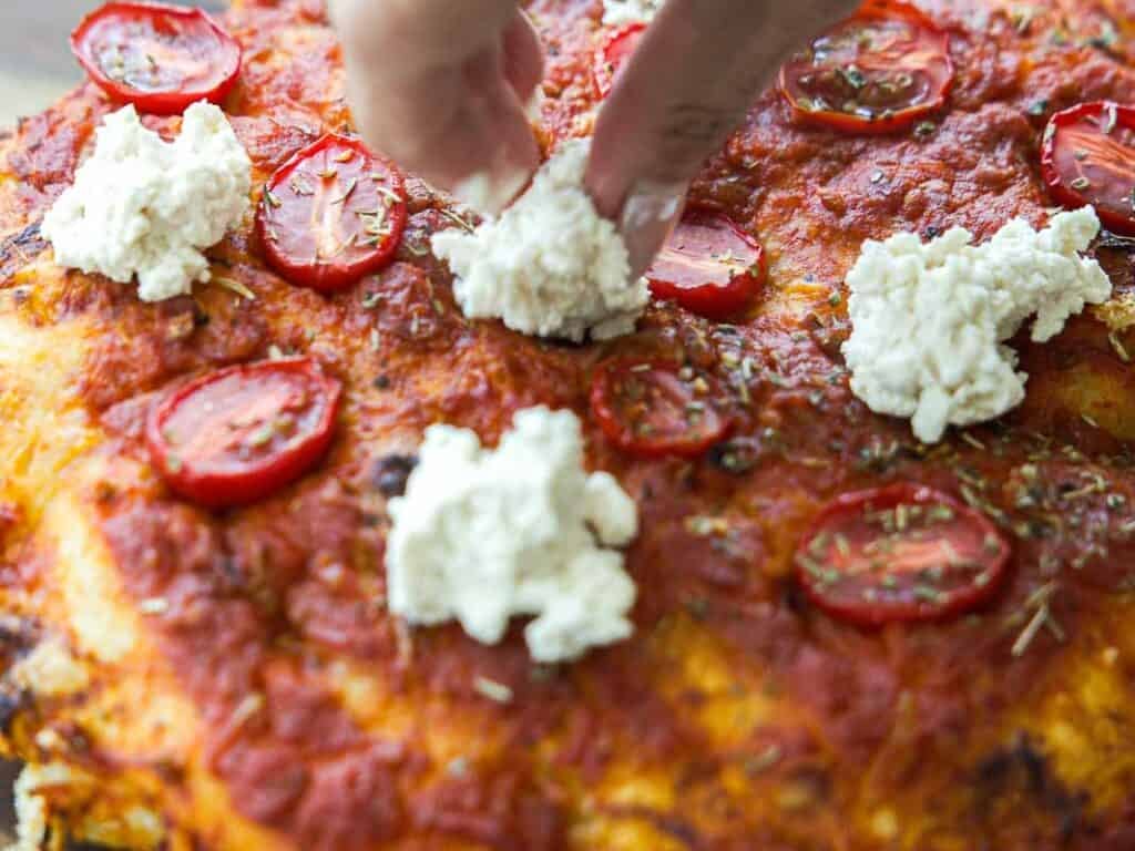 A hand sprinkles dollops of vegan ricotta cheese onto a focaccia rossa topped with tomato sauce, sliced cherry tomatoes, and herbs.