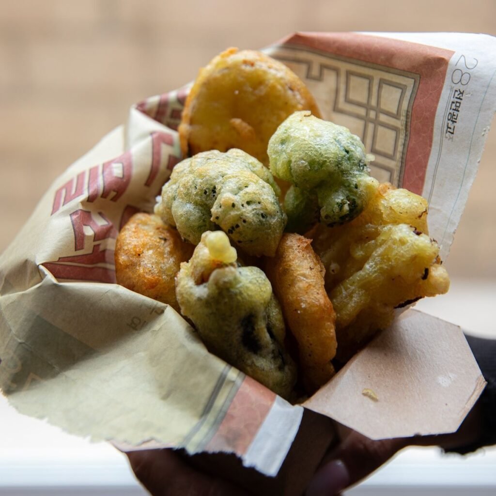 A hand holds a paper wrap filled with assorted vegetable fritters, including golden-brown and green pieces, suggesting a mix of deep-fried snacks—perfect bites to enjoy while browsing your latest Book PreOrder against a blurred background.