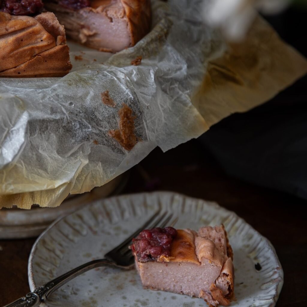 A slice of berry-topped cheesecake on a ceramic plate with a fork, in front of the larger cheesecake resting on parchment paper. The scene is dimly lit, highlighting the dessert’s creamy texture—perfect for your next Book PreOrder snack.