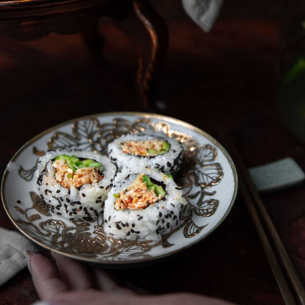 A decorative plate with three sushi rolls filled with shredded crab, avocado, and rice, sprinkled with black sesame seeds—a perfect setting to enjoy while browsing your next Book PreOrder. Chopsticks and a hand rest nearby on a dark wooden surface.
