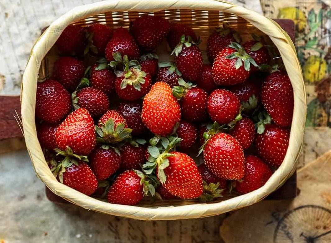 Delicious & Lemony Vegan Strawberry Shortcake Trifle. 22 A woven basket filled with fresh, ripe strawberries—perfect for making a vegan strawberry shortcake trifle—sits on a rustic surface with papers and a map partially visible underneath.