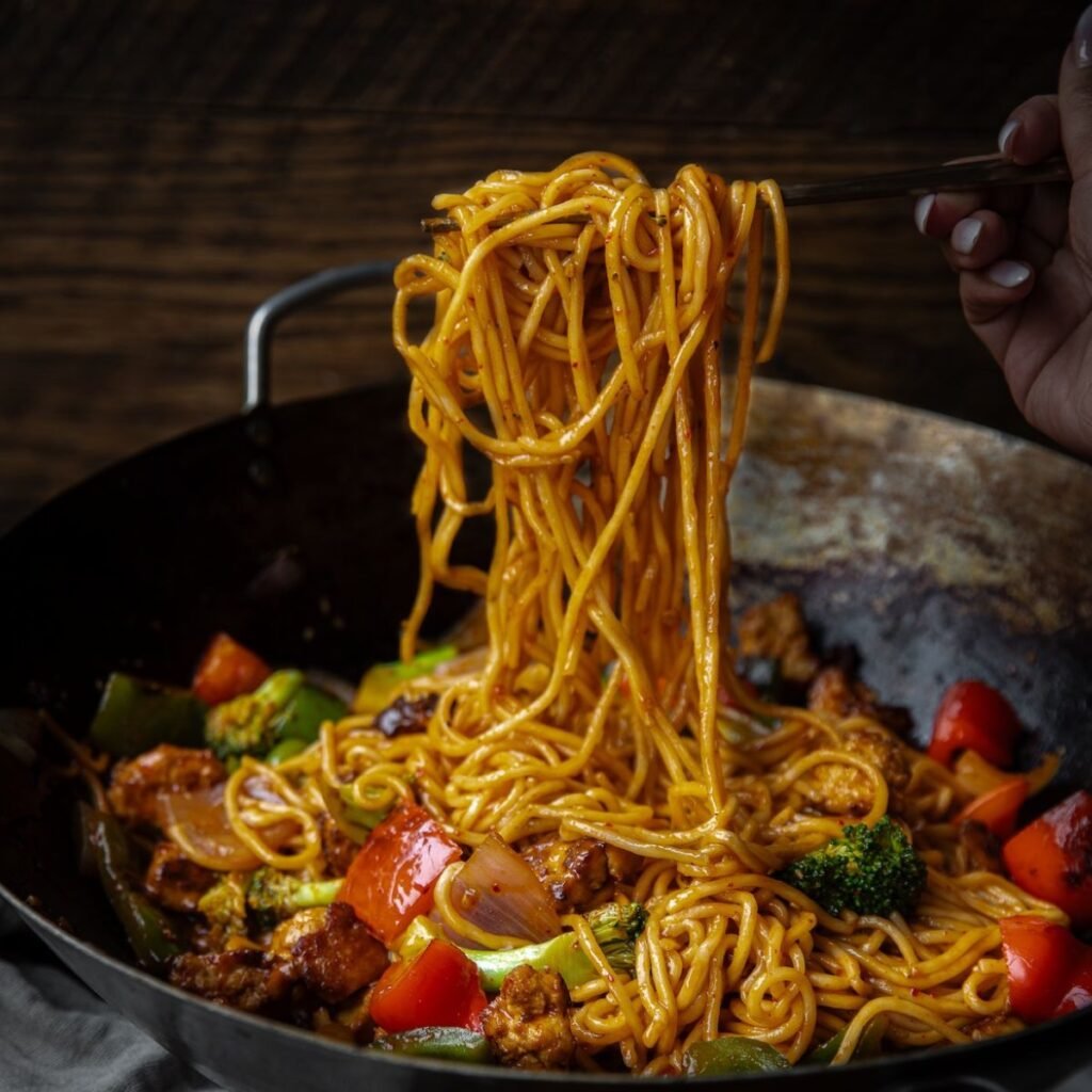 A hand holds chopsticks lifting a portion of saucy noodles from a wok filled with noodles, broccoli, bell peppers, onions, and protein—perfect fuel while browsing the Book PreOrder Test on your favorite dark wooden table.