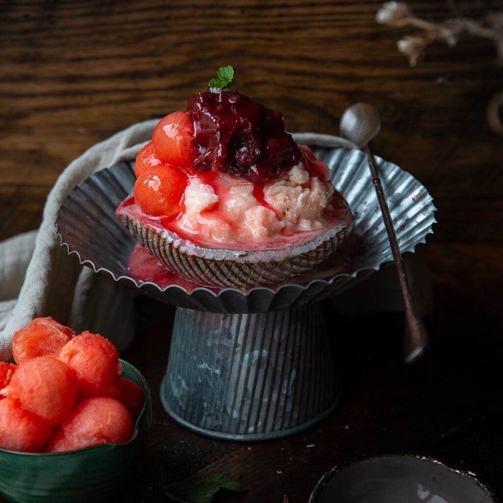 A dessert featuring scoops of pink ice cream and red melon balls, topped with berry sauce and a mint leaf, served in a shell-shaped dish with a metal spoon—a treat worth savoring while you browse your next Book PreOrder.