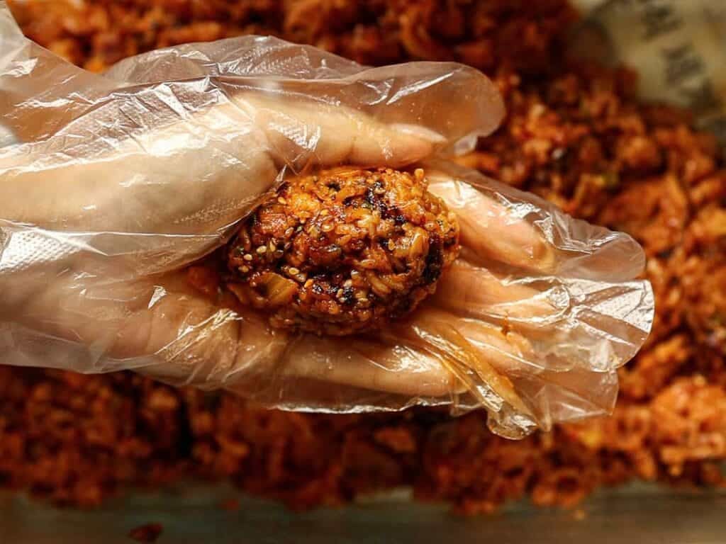 A hand wearing a clear plastic glove holds a ball of spicy, seasoned rice mixture, with more of the mixture visible in the background.