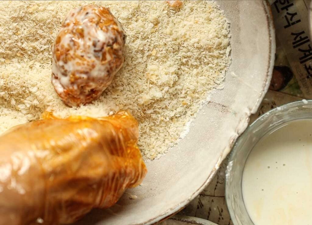A gloved hand coats a ball of food in breadcrumbs, with a bowl of batter and newspapers visible nearby.