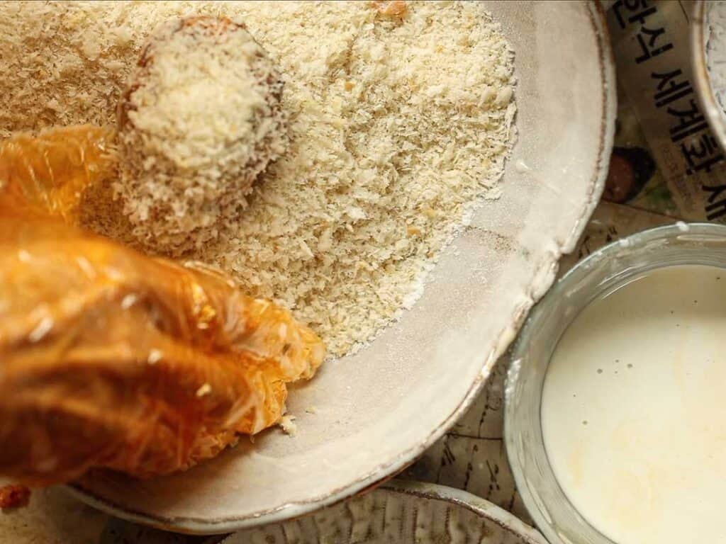 A hand in an orange glove coats food in breadcrumbs in a shallow bowl, with a bowl of liquid batter nearby.