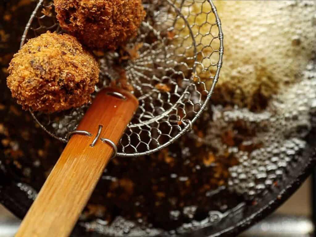 A close-up of two golden-brown, crispy fried balls being lifted from hot oil in a pan with a wire skimmer. Bubbles and oil can be seen in the background.