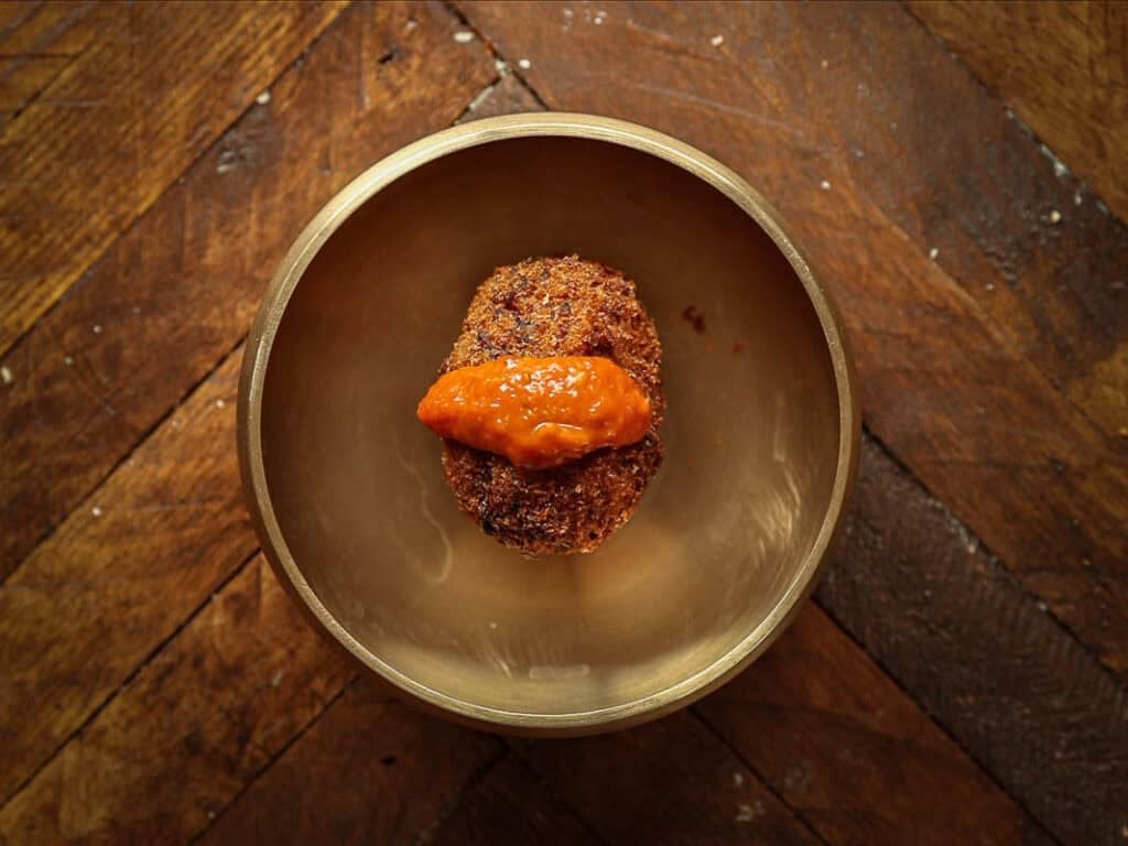 A single, round kimchi fried rice ball topped with a dollop of orange sauce sits in the center of a brass bowl on a rustic wooden table.