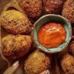 10+ Delicious and Easy Recipes Using Kimchi Today! 6 A close-up of several golden, crispy kimchi fried rice balls arranged around a bowl of bright red dipping sauce on a wooden platter. A hand with manicured nails is visible near the bottom edge of the image.