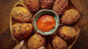 A wooden plate with several crispy, golden-brown kimchi fried rice balls arranged in a circle around a small bowl of red dipping sauce, with a hand holding the plate.