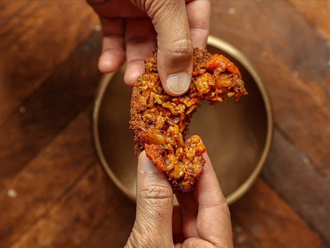 Close-up of two hands breaking apart a crispy, golden-brown kimchi fried rice ball with a visible vegetable filling over a round, empty bowl on a wooden surface.